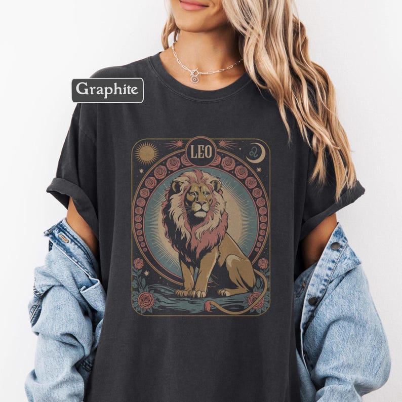 May include: Dark gray t-shirt with a graphic of a lion, sun, moon, and roses. The word "LEO" is above the lion. The shirt has the word "Graphite" in a box. The shirt is a comfortable, casual style.