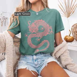 Puede incluir: Camiseta verde claro con un dise&ntilde;o de drag&oacute;n rosa. El texto "Light Green" est&aacute; en un cuadro blanco. La camiseta se combina con pantalones cortos de mezclilla y una rebeca crema. Estilo casual.