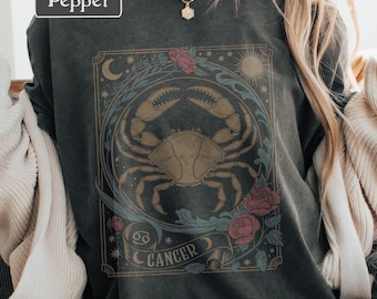 May include: Dark gray sweatshirt with a Cancer zodiac design. The design features a crab illustration in a circular frame with roses, a crescent moon, and the word "Cancer" on a banner. The word "Pepper" is in a white rectangle in the upper left corner.