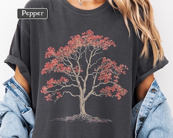 May include: Dark gray t-shirt featuring a detailed illustration of a tree with red leaves. The tree has a light brown trunk and branches, with roots visible at the base. The word "Pepper" is in a white box in the upper left corner.