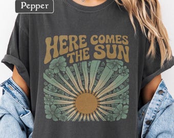May include: Charcoal grey t-shirt with the phrase "HERE COMES THE SUN" in a vintage style font. The design includes a sunburst motif with green and gold floral details. The sleeves are rolled up.