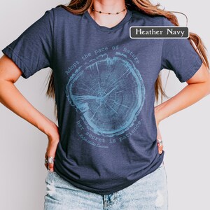 May include: Heather navy t-shirt with a tree ring graphic in light blue. The text on the shirt reads "Adopt the pace of nature, her secret is patience." - Ralph Waldo Emerson. The shirt is a crew neck.