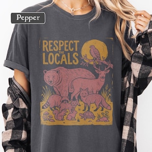 May include: Dark gray t-shirt with a graphic design featuring a bear, deer, fox, raccoon, and other animals. The text "Respect the Locals" is printed above the animals. The shirt is a Pepper brand.