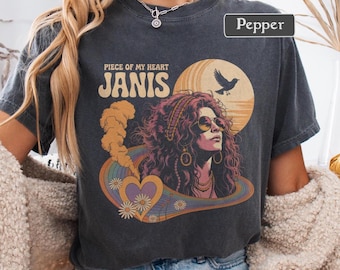 May include: Dark gray t-shirt featuring a retro graphic of Janis Joplin with the text "Piece of My Heart Janis". The design includes a rainbow, heart, and sun, evoking a vintage aesthetic. The shirt is a Pepper color.