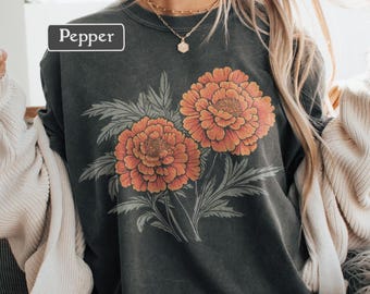 Op de afbeelding: Donkergrijze sweatshirt met een bloemenontwerp van twee oranje bloemen en groene bladeren. Het woord "Pepper" staat in een witte doos. De sweatshirt wordt door een persoon gedragen.