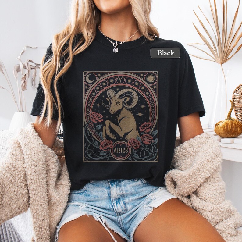May include: Black t-shirt featuring an Aries zodiac design. The graphic includes a ram, roses, and the word "ARIES" in a circular frame. The design is in shades of brown, pink, and cream.