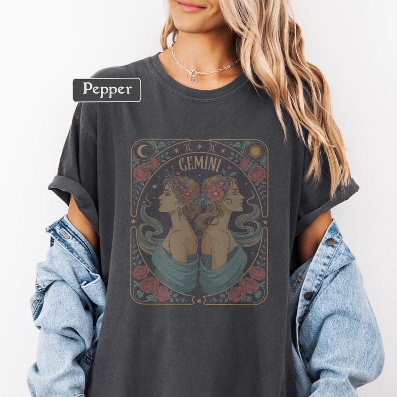 Puede incluir: Camiseta gris oscuro con un dise&ntilde;o del signo zodiacal G&eacute;minis. El gr&aacute;fico incluye dos perfiles de mujeres, flores y la palabra "Gemini" en un marco decorativo. La camiseta tiene mangas enrolladas y un corte holgado.