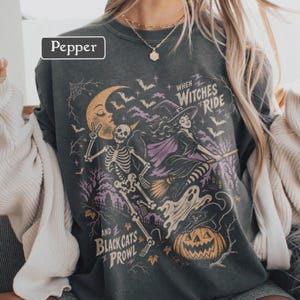 May include: A charcoal gray crewneck sweatshirt featuring a graphic print with the text "WHEN WITCHES RIDE AND BLACK CATS PROWL". The design includes a skeleton, a witch on a broomstick, a crescent moon, a ghost, a black cat, and a jack-o'-lantern.