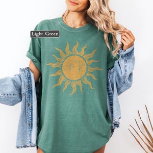 May include: Light green t-shirt with a large, distressed gold sun graphic. The shirt has short sleeves and a relaxed fit. The text "Light Green" is in a white rectangle in the upper left corner. A denim jacket is draped over the shoulders.