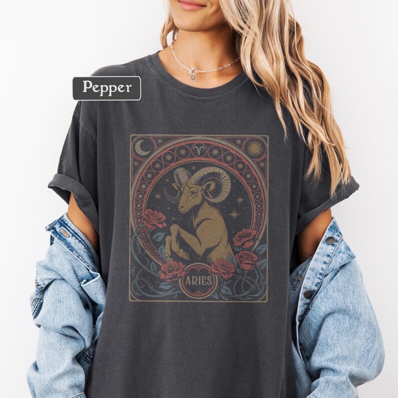 May include: Dark gray t-shirt featuring an Aries zodiac design. The graphic includes a ram, roses, and the word "ARIES" in a circular frame. The shirt has rolled-up sleeves and a relaxed fit.
