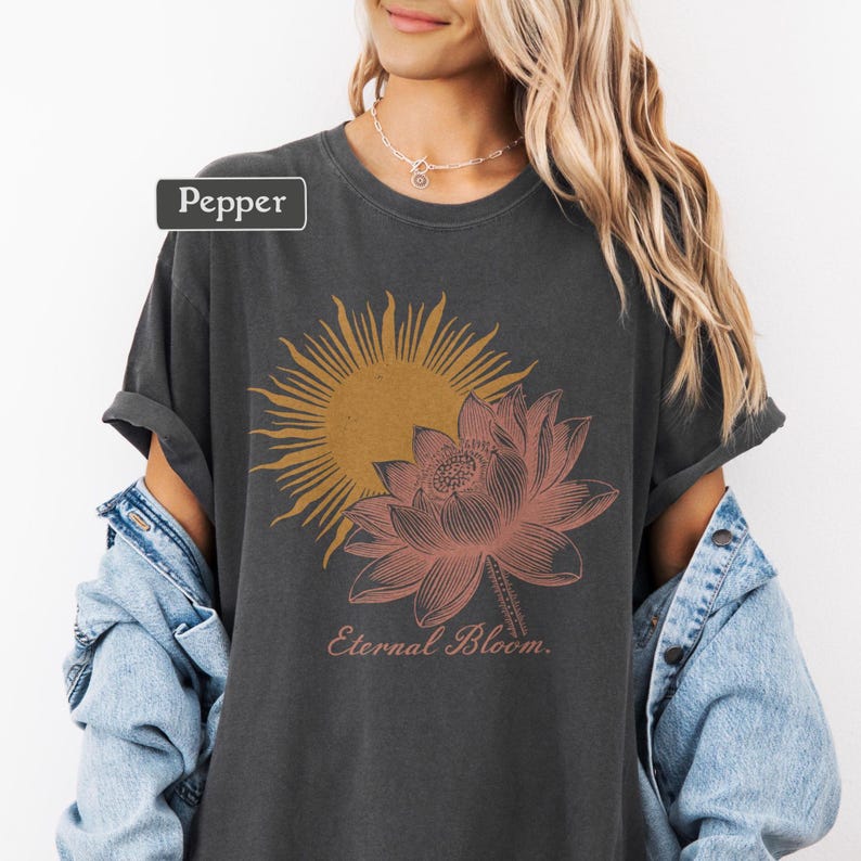 May include: Dark gray t-shirt featuring a sun and lotus flower graphic in shades of orange and pink. The text "Eternal Bloom" is below the floral design. The word "Pepper" is in a rectangular box in the upper left corner.