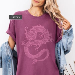 Puede incluir: Camiseta color bayas con un dise&ntilde;o detallado de drag&oacute;n rosa. La camiseta tiene mangas enrolladas y la palabra "Berry" en una etiqueta rectangular. Una chaqueta vaquera est&aacute; sobre los hombros.
