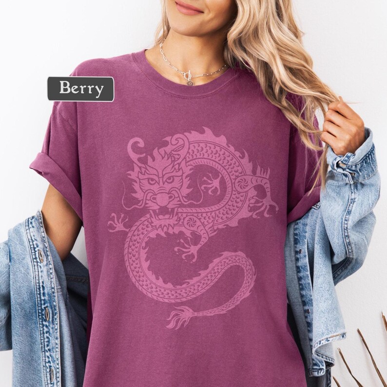 Puede incluir: Camiseta color bayas con un dise&ntilde;o detallado de drag&oacute;n rosa. La camiseta tiene mangas enrolladas y la palabra "Berry" en una etiqueta rectangular. Una chaqueta vaquera est&aacute; sobre los hombros.
