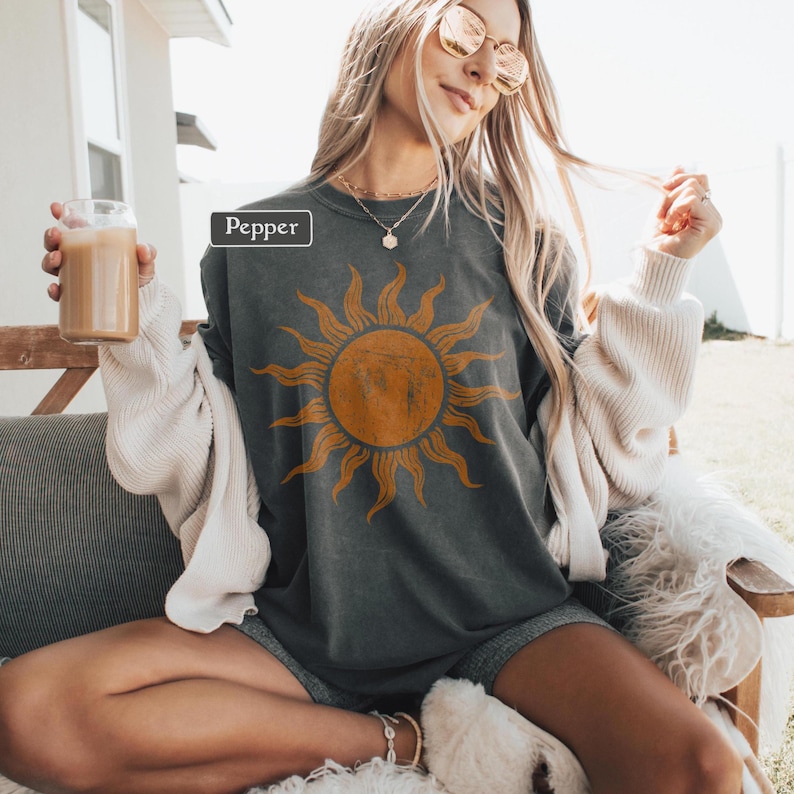 May include: Dark gray t-shirt with a large orange sun graphic. The word "Pepper" is in a white box. The shirt is worn with gray shorts and a cream-colored cardigan. A glass of a light brown beverage is held in the hand.