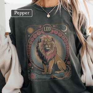 May include: Dark gray sweatshirt featuring a detailed illustration of a lion with a pink mane, surrounded by roses and celestial elements. The word "LEO" is at the top. The brand name "Pepper" is in a white box in the upper left corner.
