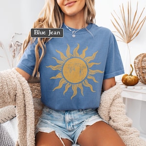 May include: Blue short-sleeved t-shirt with a distressed yellow sun graphic. The shirt has a crew neck and is a light blue color. The text "Blue Jean" is displayed in a rectangular box on the top left of the shirt.