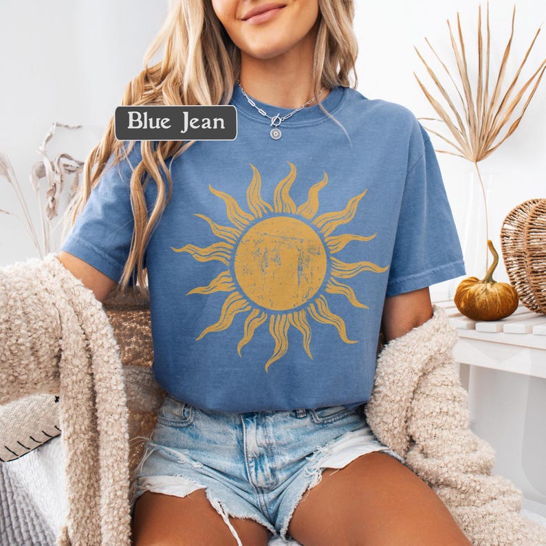 May include: Blue short-sleeved t-shirt with a distressed yellow sun graphic. The shirt has a crew neck and is a light blue color. The text "Blue Jean" is displayed in a rectangular box on the top left of the shirt.