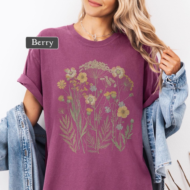 以下が含まれることがあります： ベリー色のTシャツ。黄色と緑の花柄のデザイン。袖は折り返されており、「Berry」の文字がラベルに表示されています。柔らかい素材で作られたシャツです。