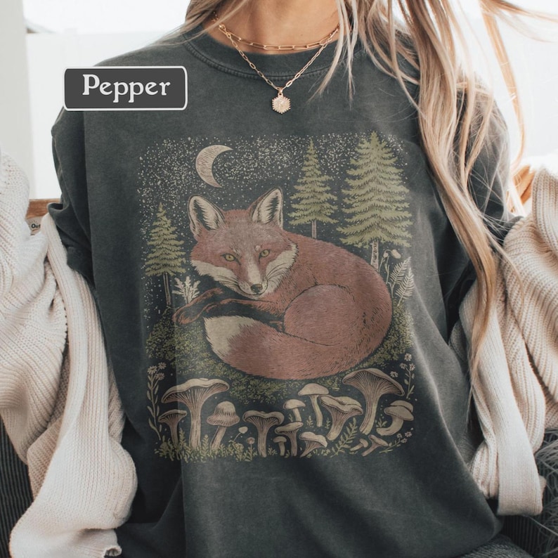 May include: Dark gray sweatshirt featuring a graphic of a curled-up fox in a forest setting with mushrooms, trees, and a crescent moon. The word "Pepper" is in a white rectangle in the upper left corner. The design is in shades of brown, green, and white.