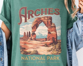 May include: Green t-shirt featuring a graphic of Arches National Park in Utah. The design includes the word "Arches" above an illustration of the park's iconic rock formations, with "National Park Utah" below. The shirt has rolled-up sleeves.