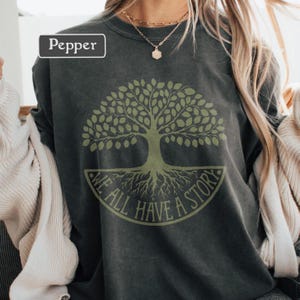 May include: Pepper-colored sweatshirt featuring a green tree graphic with the text "WE ALL HAVE A STORY" in a circular design. The sweatshirt has a crew neck and long sleeves. The word "Pepper" is in a white box in the upper left corner.