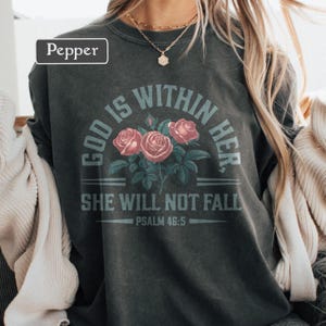 May include: Dark gray sweatshirt with the text "GOD IS WITHIN HER. SHE WILL NOT FALL. PSALM 46:5" in light blue. The design includes three pink roses with green leaves. The word "Pepper" is in a white box in the upper left corner.
