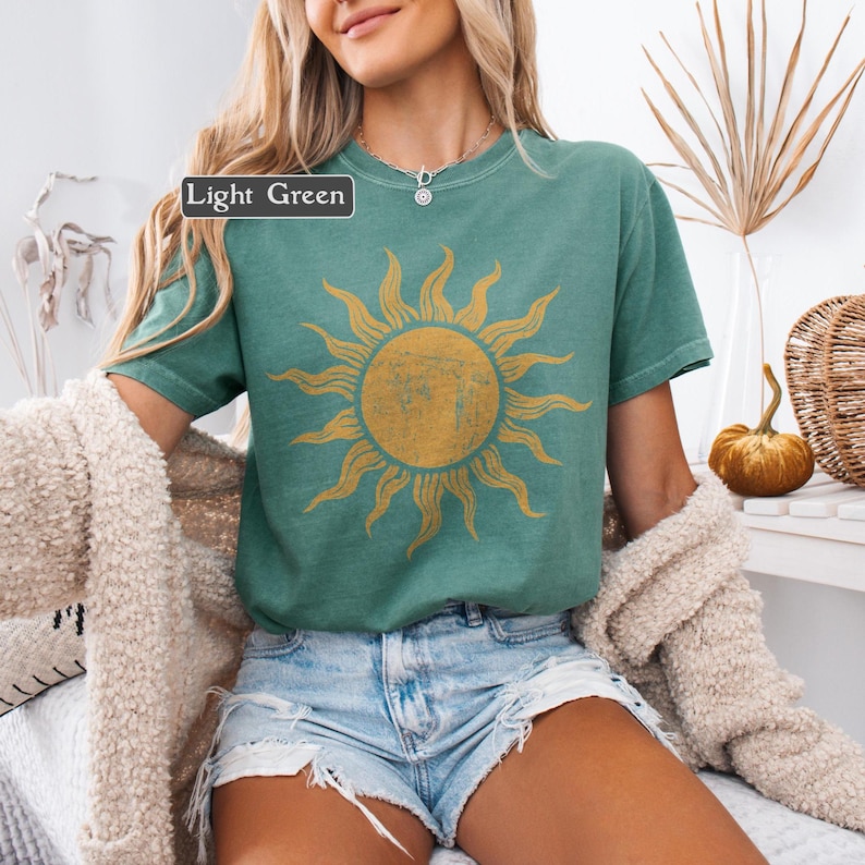 May include: Light green t-shirt featuring a distressed gold sun graphic. The shirt has short sleeves and a crew neck. The text "Light Green" is in a box in the upper left corner. The shirt is worn with denim shorts.