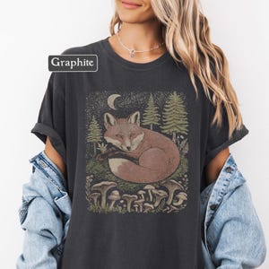May include: Graphite-colored t-shirt featuring a detailed illustration of a fox curled up in a forest scene with mushrooms, trees, and a crescent moon. The text "Graphite" is in a white box in the upper left corner.