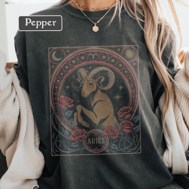 May include: Dark gray sweatshirt featuring an Aries zodiac design. The graphic includes a ram, roses, and the word "Aries" in a circular frame. The word "Pepper" is in a white rectangle in the upper left corner.