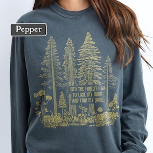 May include: A pepper-colored long-sleeve sweatshirt featuring a gold-toned graphic of a forest with pine trees and wildflowers. The text printed in the center reads: INTO THE FOREST I GO TO LOSE MY MIND AND FIND MY SOUL. The garment has a crew neckline.