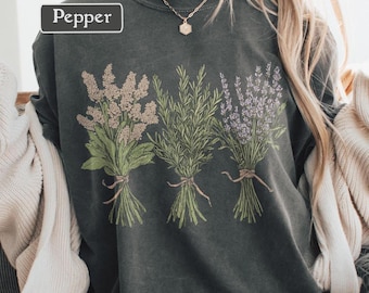 Puede incluir: Sudadera gris oscuro con una ilustración botánica de tres ramos de hierbas atados con hilo. La palabra "Pepper" está impresa en un rectángulo blanco en la esquina superior izquierda. Los ramos incluyen lavanda, romero y otras hierbas.