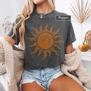 May include: A dark gray t-shirt with a distressed orange sun graphic. The word "Pepper" is in a white box on the shirt. The shirt is short-sleeved and has a vintage, worn look.