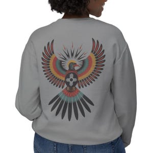 May include: A light gray sweatshirt featuring a colorful graphic of a stylized bird with outstretched wings on the back. The bird design incorporates red, yellow, blue, and black elements, with a sunburst and cross symbol.