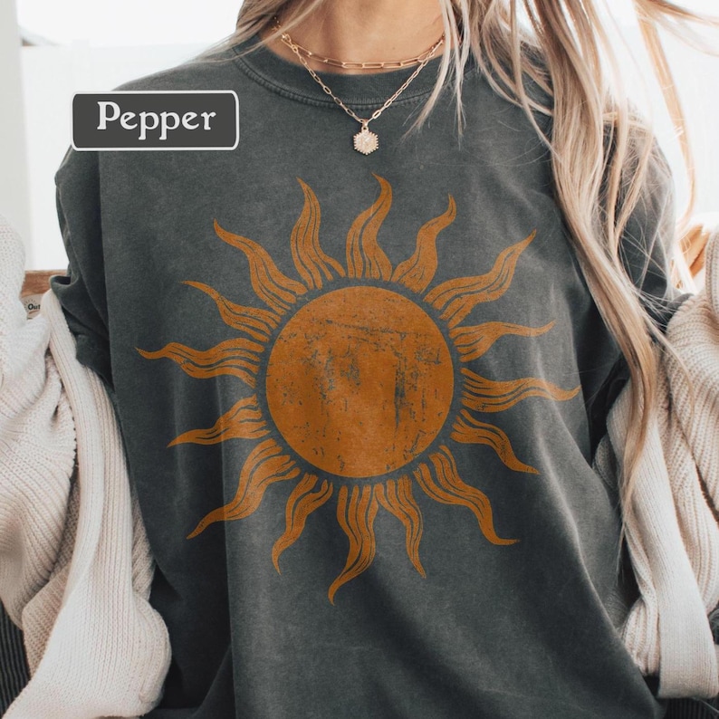 May include: A dark gray sweatshirt with a large orange sun graphic. The word "Pepper" is in a white rectangle in the upper left corner. The sweatshirt is worn with a gold necklace.