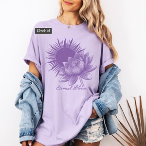 May include: Orchid-colored t-shirt featuring a sun and lotus flower design with the text "Eternal Bloom." The graphic is in a darker shade of purple. The shirt is a soft, comfortable material.