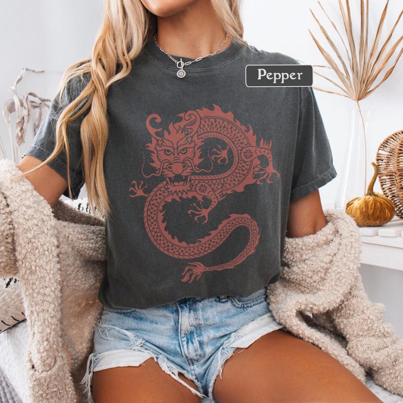 Puede incluir: Camiseta gris oscuro con un dise&ntilde;o de drag&oacute;n rojo. El dise&ntilde;o del drag&oacute;n es detallado y estilizado. La palabra "Pepper" est&aacute; en un rect&aacute;ngulo blanco. La camiseta se usa con una rebeca color crema y pantalones cortos de mezclilla.