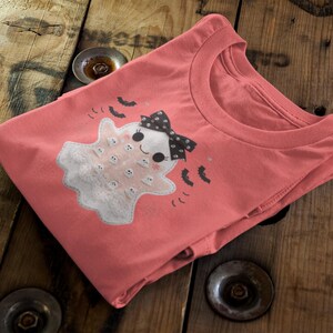 May include: A coral-colored t-shirt featuring a cute ghost design with a black bow and bats. The ghost has a white body with small white circles. The shirt is folded on a wooden surface.