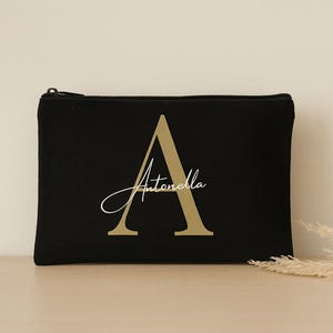 Könnte beinhalten: Schwarze Reißverschlusstasche mit goldenem Buchstaben "A" und dem Namen "Antonella" in Schreibschrift. Die rechteckige Tasche liegt auf einer hellen Oberfläche. Ein Zweig getrockneter Blumen ist im Hintergrund zu sehen.