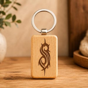 May include: A rectangular wooden keyring with a silver ring. The keyring has a dark brown engraved design on the front. The background includes a wooden surface, a vase with greenery, and a decorative item.