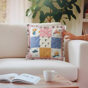 Pode incluir: Almofada quadrada de patchwork com borda rosa e quadrados coloridos. Os quadrados apresentam ilustrações temáticas do espaço, incluindo foguetes, planetas e personagens de desenhos animados. A almofada está em um sofá branco, com um livro e uma caneca em uma mesa de madeira.