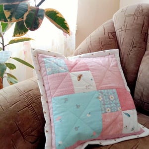 May include: A square patchwork pillow with a quilted design. The pillow features a mix of pink, light blue, and white fabric squares, some with floral and mushroom patterns. The pillow is trimmed with a white border and a pink dotted edge.