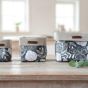 May include: Three fabric storage baskets in different sizes. They have a grey floral pattern with white details and a neutral linen-style trim. Each basket has a small brown leather tag that says "Hand Made".