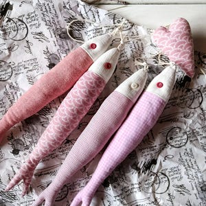 May include: Four pink fabric fish toys with white heads and red or white button eyes. The fish have different patterns, including dots, scales, and gingham. A pink heart-shaped toy is also present. The toys are arranged on patterned paper.