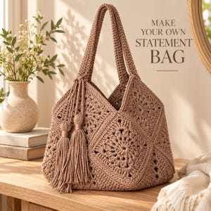 May include: A light brown crochet tote bag featuring a square motif pattern, two top handles, and two long decorative tassels hanging from one side. The text "MAKE YOUR OWN STATEMENT BAG" is printed in dark brown letters above the bag.