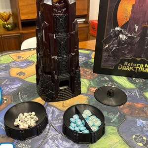 Peut inclure: Mise en place d'un jeu de société avec une tour marron foncé, deux plateaux noirs contenant des pièces de jeu et un plateau de jeu. L'illustration de la boîte de "Return to Dark Tower" est visible. Le plateau de jeu a un design de carte coloré.