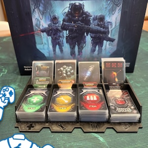 May include: A board game with a box featuring soldiers in futuristic armor. The game includes cards with various icons and text, such as "ITEM" and "SERIOUS WOUND". The cards are organized in a black plastic tray.
