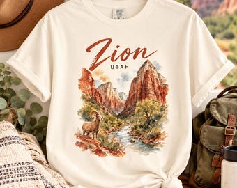 May include: A cream-coloured short-sleeve t-shirt featuring a watercolour-style graphic of Zion National Park in Utah. The design includes red rock cliffs, a flowing river, green vegetation, and a bighorn sheep, with the text Zion UTAH printed above.