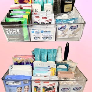May include: Two clear plastic rectangular storage bins filled with various travel-sized personal care items, including Dude Wipes, Purell hand sanitizer packets, Shout stain remover wipes, TRESemmé hair gel, and lens cleaning wipes in organized sections.