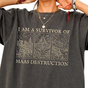 May include: Dark gray t-shirt with the text "I AM A SURVIVOR OF MAAS DESTRUCTION" and three illustrated panels. The panels depict cityscapes and castles. A 100% cotton tag is visible.