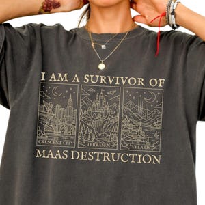 以下が含まれることがあります： チャコールグレーの半袖Tシャツ。都市や風景を描いた3つの長方形パネルのグラフィックプリントが施されています。テキストには「I AM A SURVIVOR OF MAAS DESTRUCTION」と記され、Crescent City、Terrasen、Velarisのラベルが付いています。